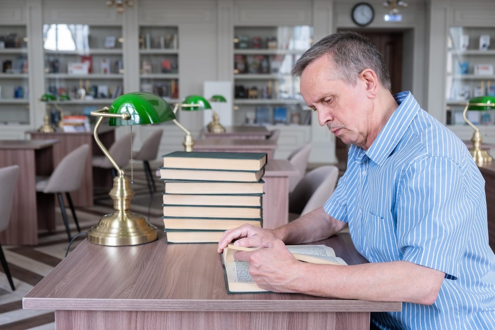 Homem lendo atentamente em uma escrivaninha de biblioteca, com uma pilha de livros e um abajur de latão ao lado